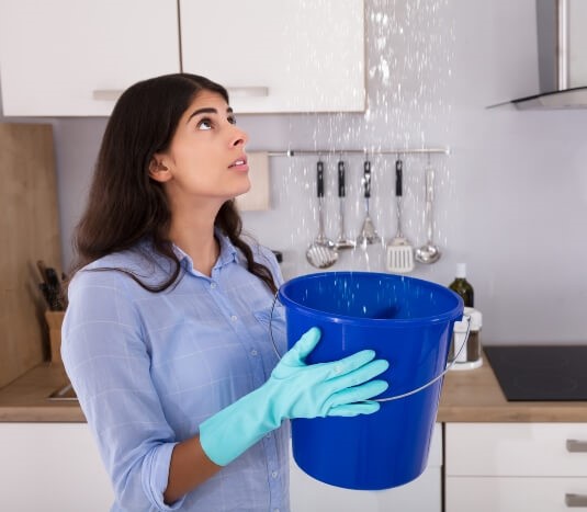 Woman holding a bucket under a leaking roof.
